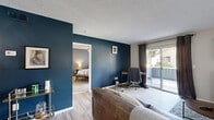 This well-appointed single-story residence in Escondido, California, offers a thoughtful layout across five distinct rooms, making efficient use of every square foot. The living room features a textured ceiling, plush carpeting, and wall space ideal for a mounted TV, lending a cozy yet functional atmosphere. Adjacent to it, the living area extends to a room with light wood-look flooring, a bold navy accent wall, and a sliding glass door that floods the space with natural light and provides outdoor access. The galley-style kitchen is lined with crisp white cabinetry, grey countertops, and stainless steel appliances, including a refrigerator and range, all illuminated by a skylight overhead. The dedicated dining area features wood-look flooring and a ceiling fan with light fixture, creating a pleasant space for meals. The bedroom is a comfortable retreat, highlighted by a navy accent wall, soft carpeting, a ceiling fan, and a window dressed with grey curtains that allow natural light to filter through. The bathroom is clean and functional, offering a vanity with grey countertop, a large mirror, and a shower-tub combination. Throughout the home, consistent neutral tones and modern finishes create a cohesive, inviting ambiance.