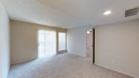This single-story space presents a straightforward and functional layout centered around a generously sized main living area. The room features wall-to-wall neutral-toned carpeting that adds warmth and comfort underfoot, complemented by smooth beige walls and a textured ceiling that together create a cohesive, understated aesthetic. A large sliding glass door dominates one wall, fitted with vertical blinds that allow for adjustable natural light control. Alongside it, a single window with a horizontal blind provides an additional source of daylight, brightening the interior. Recessed lighting is visible near the entryway, supplementing natural light as needed. The open wall space offers flexibility for furniture arrangement throughout the room. An open doorway leads to an adjoining room, also carpeted, with a ceiling-mounted light fixture visible, suggesting it may serve as a bedroom or secondary living space. Clean white baseboards frame the perimeter of the room, adding a crisp finishing detail. Overall, the space presents a clean, move-in ready environment with a neutral palette suited to a variety of interior styles.