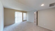This single-story space presents a straightforward and functional layout centered around a generously sized main living area. The room features wall-to-wall neutral-toned carpeting that adds warmth and comfort underfoot, complemented by smooth beige walls and a textured ceiling that together create a cohesive, understated aesthetic. A large sliding glass door dominates one wall, fitted with vertical blinds that allow for adjustable natural light control. Alongside it, a single window with a horizontal blind provides an additional source of daylight, brightening the interior. Recessed lighting is visible near the entryway, supplementing natural light as needed. The open wall space offers flexibility for furniture arrangement throughout the room. An open doorway leads to an adjoining room, also carpeted, with a ceiling-mounted light fixture visible, suggesting it may serve as a bedroom or secondary living space. Clean white baseboards frame the perimeter of the room, adding a crisp finishing detail. Overall, the space presents a clean, move-in ready environment with a neutral palette suited to a variety of interior styles.