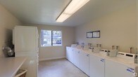 This single-floor shared laundry facility is designed for efficiency and ease of use. The room is clean and functional, featuring a lineup of white top-load washing machines arranged along one wall, paired with a matching row of dryers to accommodate multiple users simultaneously. A stacked set of machines is also positioned along the adjacent wall, maximizing the room's capacity. The space is illuminated by bright overhead fluorescent lighting, ensuring excellent visibility throughout. A window on the far wall allows natural light to filter in while offering a pleasant view of the surrounding outdoor greenery, contributing to an open and airy atmosphere. The walls are finished in a neutral light tone, complementing the clean, polished concrete-style flooring that is both durable and easy to maintain. Informational signage is posted on the wall for user reference, providing instructions and guidelines for operating the machines. Overall, the facility is thoughtfully organized to serve a communal setting, offering a practical and well-maintained environment for laundry needs.