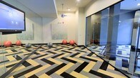 This contemporary fitness studio is designed with both functionality and aesthetic appeal in mind. The space immediately draws the eye with its striking geometric-patterned carpet tiles, composed of interlocking black, grey, and golden-yellow chevrons that create a dynamic visual foundation across the floor. Full-length mirrored wall panels line one side of the room, amplifying the sense of space and allowing users to monitor their form during workouts. A collection of exercise balls in varying sizes — grey, red, and pink — are stationed along the base of the mirrored wall, ready for use. A piece of abstract wall art featuring muted green tones adds a calming, decorative touch to the otherwise energetic environment. Overhead, a suspended disco ball pendant light serves as a playful yet stylish focal point, catching and reflecting light throughout the room. A glass partition wall with a sliding door separates an adjacent section of the studio, offering a degree of privacy while maintaining an open, connected feel. The neutral wall tones and recessed ceiling details contribute to a clean, modern finish that keeps the space feeling bright and welcoming. Overall, the studio strikes a balance between professional fitness utility and contemporary interior design.