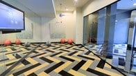 This contemporary fitness studio is designed with both functionality and aesthetic appeal in mind. The space immediately draws the eye with its striking geometric-patterned carpet tiles, composed of interlocking black, grey, and golden-yellow chevrons that create a dynamic visual foundation across the floor. Full-length mirrored wall panels line one side of the room, amplifying the sense of space and allowing users to monitor their form during workouts. A collection of exercise balls in varying sizes — grey, red, and pink — are stationed along the base of the mirrored wall, ready for use. A piece of abstract wall art featuring muted green tones adds a calming, decorative touch to the otherwise energetic environment. Overhead, a suspended disco ball pendant light serves as a playful yet stylish focal point, catching and reflecting light throughout the room. A glass partition wall with a sliding door separates an adjacent section of the studio, offering a degree of privacy while maintaining an open, connected feel. The neutral wall tones and recessed ceiling details contribute to a clean, modern finish that keeps the space feeling bright and welcoming. Overall, the studio strikes a balance between professional fitness utility and contemporary interior design.