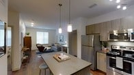 This contemporary single-floor residence showcases a thoughtfully designed open-concept layout that seamlessly connects the kitchen, dining, and living areas into one cohesive living space. The kitchen is a standout feature, anchored by a spacious island with a sleek countertop and bar seating, making it ideal for casual dining or entertaining. Warm wood-toned cabinetry pairs elegantly with stainless steel appliances, including a full-size refrigerator, while stylish pendant lights hang above the island, adding a modern touch to the space. Durable hardwood-style flooring runs throughout, lending warmth and continuity across the open floor plan. The living area is bathed in natural light from a large window, creating a bright and welcoming atmosphere. The neutral color palette, featuring crisp white walls and earthy accents, contributes to a clean, modern aesthetic. A dedicated workspace or dining nook is visible in the background, adding to the unit's functionality. The overall ambiance is contemporary and inviting, blending practical design with stylish finishes to create a comfortable and well-appointed living environment.