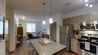 This contemporary single-floor residence showcases a thoughtfully designed open-concept layout that seamlessly connects the kitchen, dining, and living areas into one cohesive living space. The kitchen is a standout feature, anchored by a spacious island with a sleek countertop and bar seating, making it ideal for casual dining or entertaining. Warm wood-toned cabinetry pairs elegantly with stainless steel appliances, including a full-size refrigerator, while stylish pendant lights hang above the island, adding a modern touch to the space. Durable hardwood-style flooring runs throughout, lending warmth and continuity across the open floor plan. The living area is bathed in natural light from a large window, creating a bright and welcoming atmosphere. The neutral color palette, featuring crisp white walls and earthy accents, contributes to a clean, modern aesthetic. A dedicated workspace or dining nook is visible in the background, adding to the unit's functionality. The overall ambiance is contemporary and inviting, blending practical design with stylish finishes to create a comfortable and well-appointed living environment.