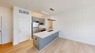 Located at 500 Maury St, this single-floor unit offers a thoughtfully designed layout across four functional spaces. The contemporary kitchen serves as a central highlight, featuring a large island with a built-in sink, crisp white quartz countertops, gray cabinetry, and a full suite of stainless steel appliances including a refrigerator, electric range, and built-in microwave. A sleek pendant light fixture anchors the island, while light-toned hardwood flooring flows seamlessly throughout the living areas, enhancing the bright and airy atmosphere. The bedroom is generously appointed with two dedicated closets offering ample built-in shelving and hanging space, along with a ceiling fan and a window that invites natural light. The full bathroom includes a soaking tub with shower, a vanity, and clean tile flooring in a neutral palette. Throughout the unit, white walls, clean lines, and modern finishes create a cohesive, minimalist aesthetic. The open sightlines from the kitchen through the hallway contribute to an efficient and well-connected flow, making the space feel open and functional despite its compact footprint.