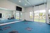 Located at 289 Lakeshore Pkwy in Birmingham, Alabama, this single-story fitness center offers a well-appointed space designed to support a variety of exercise routines. The room is finished with teal carpeted flooring accented by a diamond-patterned tile inlay, providing a visually engaging and cushioned surface suitable for both equipment use and floor-based workouts.

A standout architectural feature is the full-width glass garage door along one wall, flanked by additional windows that flood the interior with abundant natural light and provide unobstructed views of the surrounding green landscaped grounds. This open, airy quality is further enhanced by the elevated ceilings and recessed lighting throughout.

The space is equipped with cardio machines, including an elliptical trainer and a WaterRower Club rowing machine. A wall-mounted pull-up bar adds functional training options. One wall features a large mirror, useful for monitoring form during exercise. A fitness rules placard is posted near the entryway, indicating shared-use protocols for the space.

Multiple doors provide access to adjacent areas, including what appears to be a restroom. The clean, neutral wall tones and bright finishes contribute to an inviting and motivating atmosphere well-suited for daily fitness use.
