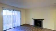 This single-floor space presents a clean and functional layout with a welcoming living area at its heart. The room features rich, dark hardwood-style flooring that grounds the space and contrasts neatly with the light-toned walls, giving the interior a balanced and neutral aesthetic. A standout element is the fireplace, framed by a dark granite surround and mantel that adds a touch of architectural character and serves as a natural focal point within the room. Adjacent to the fireplace, a large sliding glass door dressed with vertical blinds floods the space with natural light, creating a bright and open atmosphere. The door provides access to the outdoors, further enhancing the sense of connectivity and airiness. The ceiling features a subtle textured finish consistent throughout the space. The overall ambiance is understated and versatile, offering a solid foundation that can be adapted to a range of interior styles. The absence of clutter and the neutral color palette make the architectural and design details of the space stand out clearly, highlighting the quality of the finishes and the thoughtful layout of the living area.