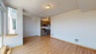 This single-floor unit offers a clean, modern layout designed for comfortable everyday living. The open living area is finished with warm, light-toned hardwood-style flooring that runs throughout, complemented by crisp white walls and wood-toned baseboards that lend a cohesive aesthetic across the space.

The kitchen is efficiently designed and positioned at the edge of the main living area. It features medium-toned wood cabinetry with sleek bar-pull hardware, light-colored countertops, and a double stainless steel sink with a pull-down faucet. Stainless steel appliances include a range with electric cooktop, an over-the-range microwave, and a dishwasher, providing a fully functional cooking environment.

The bathroom features a matching wood-toned vanity with ample cabinet storage, a light countertop, an integrated sink, and a large wall mirror that enhances the sense of space. The flooring continues the hardwood-style theme seen throughout the unit.

A separate room, accessible via a sliding barn-style door, offers flexible use as a bedroom or additional living space, with ample wall space and consistent flooring. Large windows in one of the rooms allow generous natural light to fill the interior, offering views of the surrounding outdoor landscape and contributing to an airy, open ambiance throughout the unit.