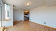 This single-floor unit offers a clean, modern layout designed for comfortable everyday living. The open living area is finished with warm, light-toned hardwood-style flooring that runs throughout, complemented by crisp white walls and wood-toned baseboards that lend a cohesive aesthetic across the space.

The kitchen is efficiently designed and positioned at the edge of the main living area. It features medium-toned wood cabinetry with sleek bar-pull hardware, light-colored countertops, and a double stainless steel sink with a pull-down faucet. Stainless steel appliances include a range with electric cooktop, an over-the-range microwave, and a dishwasher, providing a fully functional cooking environment.

The bathroom features a matching wood-toned vanity with ample cabinet storage, a light countertop, an integrated sink, and a large wall mirror that enhances the sense of space. The flooring continues the hardwood-style theme seen throughout the unit.

A separate room, accessible via a sliding barn-style door, offers flexible use as a bedroom or additional living space, with ample wall space and consistent flooring. Large windows in one of the rooms allow generous natural light to fill the interior, offering views of the surrounding outdoor landscape and contributing to an airy, open ambiance throughout the unit.