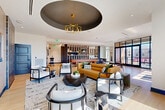 Located at 1350 Dresden Dr NE in Brookhaven, Georgia, this contemporary single-floor space offers a thoughtfully designed layout encompassing a living room, kitchen, and balcony. The living area is a standout, anchored by a striking circular ceiling medallion with a bold geometric gold chandelier. Light hardwood flooring runs throughout, complementing the bright, open atmosphere created by floor-to-ceiling black-framed glass walls and windows that flood the interior with natural light and frame views of the surrounding Brookhaven neighborhood. Seating areas are arranged to encourage both conversation and relaxation, with a warm-toned sofa, contemporary accent chairs, and a curated mix of furnishings that blend modern and organic elements. A dedicated dining area with a round table sits adjacent to the expansive glazed façade. The kitchen is equally impressive, featuring an extra-long island with a sleek white quartz countertop, light wood cabinetry with matte black hardware, a dramatic textured stone accent wall, and a cascading pendant light fixture that runs the full length of the island. Stainless steel appliances complete the professional-quality setup. The balcony extends the living space outdoors, offering open-air views of the vibrant Brookhaven area.