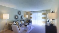 This single-floor space offers a comfortable and stylishly designed interior that blends functionality with modern aesthetics. The living area serves as the focal point of the space, featuring plush wall-to-wall carpeting that adds warmth underfoot. A large sliding glass door floods the room with an abundance of natural light, complemented by bold gold and white striped draping curtains that frame the view of the outdoors. Vertical blinds provide flexible light control throughout the day. The living room is finished with a textured ceiling and neutral-toned walls that create a calm, inviting atmosphere. Thoughtful décor details, including a set of decorative sunburst wall mirrors, contribute a touch of artistic character to the space. The seating arrangement centers around a plush sectional sofa accented with an array of patterned and solid throw pillows in grey and gold tones, accompanied by a sleek, light-toned coffee table. The overall ambiance is cohesive and contemporary, with a neutral color palette punctuated by gold accents that lend a sense of warmth and sophistication to the interior.