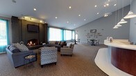 This single-floor space offers a well-appointed communal living area designed for both relaxation and social gathering. The room is generously sized, with a warm and inviting atmosphere anchored by a floor-to-ceiling brick fireplace on one wall, accompanied by a wall-mounted flat-screen television. Recessed lighting is distributed across the ceiling, providing consistent illumination throughout, while large windows dressed with teal accent curtains allow ample natural light to fill the space.

The lounge area features a comfortable arrangement of sofas and patterned accent chairs centered around the fireplace, creating an ideal setting for casual conversation or quiet relaxation. The carpeted flooring in a neutral tone adds warmth and cohesion to the overall design. Toward the back of the room, a dedicated dining area is furnished with bistro-style tables and chairs, offering a more structured setting for meals or casual meetings.

The walls are painted in a soft grey tone, accented with framed artwork and decorative shelving, contributing to the space's modern yet approachable aesthetic. The open layout and thoughtful furnishing make this a versatile and welcoming environment.