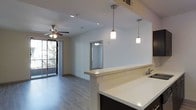This single-floor space showcases a contemporary open-concept design that seamlessly connects the kitchen and living areas. The kitchen features a clean white countertop peninsula that serves as a natural divider, complemented by stylish cylindrical pendant lights suspended from the ceiling. Dark cabinetry beneath the peninsula provides a striking contrast to the light countertop surfaces.

The living area is finished with smooth hardwood-style flooring throughout, lending warmth and continuity to the space. A ceiling fan with integrated lighting is centrally positioned, providing both comfort and ambient illumination. Recessed lighting throughout further enhances the bright, airy atmosphere.

Large sliding glass doors lead to an outdoor balcony, allowing natural light to flood the interior while offering views of surrounding greenery. A doorway visible in the background hints at additional rooms beyond the main living space.

The overall aesthetic is clean and modern, with neutral white walls, gray-toned flooring, and carefully chosen fixtures that contribute to a cohesive, well-appointed interior. The thoughtful layout maximizes the sense of openness while maintaining functional distinctions between living and kitchen zones.
