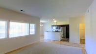 This single-floor space offers a clean, contemporary design characterized by an open-concept layout that connects the living area and kitchen in a seamless, flowing arrangement. The living area features light-toned carpeting and crisp white walls, creating a bright and welcoming atmosphere. Natural light pours in through large windows dressed with horizontal blinds, enhancing the airy feel of the space.

The kitchen is a standout feature, equipped with modern stainless steel appliances including a refrigerator, microwave, and range. Rich dark cabinetry provides a striking contrast against the lighter tones of the surrounding space, while a peninsula with a dark countertop offers additional workspace and a natural division between the kitchen and living areas. Recessed lighting and a decorative chandelier above the peninsula add both function and a touch of elegance to the kitchen zone.

The transition between the carpeted living area and the hard flooring of the kitchen creates a natural delineation between the two spaces while maintaining the open, cohesive feel of the overall layout. Neutral tones throughout contribute to a timeless and versatile aesthetic, making this a highly functional and visually appealing space.
