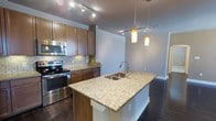 This well-appointed single-floor residence offers three bedrooms and two bathrooms, delivering a comfortable and functional layout. The heart of the home is the open-concept kitchen, which features rich wood cabinetry, a decorative tile backsplash, and gleaming granite countertops that extend across a spacious central island with a double undermount sink. Stainless steel appliances, including a range and microwave, add a modern, polished touch. Pendant lights hang above the island, complementing the track lighting overhead to create a warm, well-lit cooking and entertaining space. Dark hardwood flooring flows throughout, providing a sophisticated contrast to the light-toned walls and countertops. The open sightlines from the kitchen into the adjacent living area promote a seamless, connected feel ideal for both everyday living and hosting. The three bedrooms offer dedicated private retreats, while the two bathrooms provide convenience for residents and guests alike. With its thoughtful design, quality materials, and cohesive aesthetic, this residence presents an inviting and practical living environment suited for a variety of lifestyles.