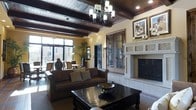 This single-floor space showcases a striking Spanish Colonial-inspired aesthetic, blending warm tones, rich textures, and carefully curated architectural details throughout. The main living area commands attention with its dramatic dark wood ceiling beams and planked wood ceiling, lending a sense of grandeur and warmth. A wrought-iron chandelier serves as a centerpiece overhead, casting ambient light across the generously proportioned room.

A substantial stone fireplace with an ornately detailed surround anchors one wall, providing both visual and functional appeal. Flanking it are elegant double doors crafted from dark wood and intricate wrought-iron scrollwork, adding a refined, old-world character to the space. Warm golden-hued walls complement the dark wood furnishings, including a rustic console cabinet and heavy coffee table, creating a cohesive and inviting palette.

Large windows along one side flood the interior with natural light and offer views to an outdoor area, enhancing the openness of the space. Lush indoor plants add a touch of greenery, softening the bold architectural elements. A separate dining or bar area is visible beyond decorative columns, fitted with high seating and pendant lighting. Decorative accents, including hand-painted ceramic vessels and wall art, complete the sophisticated, character-rich atmosphere of this distinctive space.