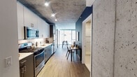 Located at 60 E Randolph Street in the heart of Chicago's Loop, this single-floor residence showcases a compelling blend of industrial and contemporary design. Exposed concrete ceilings run throughout the space, creating a striking architectural contrast against clean white walls and warm wood-tone flooring.

The kitchen is thoughtfully designed with a galley-style layout, featuring two-tone cabinetry — white uppers and gray lowers — paired with white quartz countertops, a subway tile backsplash, and a full suite of stainless steel appliances including a gas range, microwave, dishwasher, and refrigerator. Track lighting illuminates the workspace efficiently.

The living area offers an open, comfortable layout with room for a full seating arrangement. The exposed concrete ceiling continues here, adding texture and urban character to the space. A dedicated dining area near the large windows allows for natural light to fill the room throughout the day.

The bedroom features floor-to-ceiling windows with views of the surrounding cityscape, bringing in ample natural light while maintaining a calm, restful atmosphere. The bathroom is finished with clean, modern fixtures, including a glass-enclosed walk-in shower, a dark-toned vanity with white countertop, and white tile flooring. The overall ambiance is refined, urban, and functional — well-suited for life in the heart of Chicago.