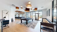 Located at 901 E Lincoln Hwy in Exton, PA, this generously sized single-floor clubhouse delivers an upscale community amenity experience with thoughtfully designed spaces flowing seamlessly into one another. The main living and entertainment area is a showstopper, anchored by a full-size pool table set beneath an artful arrangement of drum pendant lighting. Large floor-to-ceiling black-framed glass doors open directly to an outdoor terrace, flooding the interior with natural light and offering sweeping views of the surrounding landscape. A dramatic herringbone-patterned hardwood floor adds texture and warmth throughout the living and dining zones. The dining area features an impressive live-edge communal table beneath soaring vaulted ceilings with exposed wood beams and linear pendant fixtures. Adjacent to the entry, a stylish wet bar with a quartz waterfall countertop, mosaic tile surround, and leather bar seating provides an elegant gathering point near a stainless steel elevator. The fitness center is well-appointed with cardio equipment, cycling stations, and a striking topographic mural accent wall, all finished with the same herringbone hardwood flooring. A curved glass-railed mezzanine overlooks the lower level, adding a refined architectural element. The overall ambiance is modern, warm, and inviting.