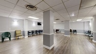 This single-floor commercial space offers a spacious, open layout well-suited for professional or clinical operations. The interior is defined by clean, neutral tones, with walls finished in a two-tone gray scheme divided by white chair-rail molding — a detail that lends a polished, organized character throughout. Light wood-look vinyl flooring runs continuously across the space, contributing to a bright and cohesive aesthetic.

The main area is expansive and open, supported by structural columns finished to match the surrounding walls. Seating is arranged throughout in multiple configurations — rows of modern chairs along the perimeter walls and small grouped seating clusters — suggesting dedicated waiting zones. Wall-mounted sconce lighting provides warm accent illumination, complementing the recessed and panel lighting integrated into the suspended drop-tile ceiling, which also features circular HVAC diffusers for climate control.

A wall-mounted flat-screen television is positioned in the primary waiting area, enhancing the comfort and functionality of the space. A secondary area, partially separated by a half-wall partition, provides a more intimate seating arrangement ideal for smaller gatherings or consultations.

A compact kitchenette is also present, outfitted with dark espresso cabinetry, stone-patterned countertops, a stainless steel sink, and a microwave — offering convenient on-site utility for staff or occupants. Overall, the space presents a clean, professional environment ready for immediate use.