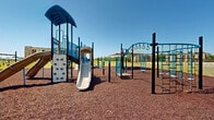Located at 1151 GA Highway 96 in Kathleen, Georgia, this outdoor recreational playground serves as a central amenity within a multi-building residential community. The play area is expansively laid out on a rubberized mulch surface, providing a safe and cushioned ground cover throughout. Multiple play structures are distributed across the space, including climbing towers with rock walls, dual-lane slides, and tunnel elements, all finished in a distinctive blue and grey color scheme. A dedicated swing set offers additional play options alongside the main structures. A large shade sail canopy anchors one section of the playground, providing a covered retreat from the Georgia sun and creating a comfortable gathering point. Curved brick edging neatly defines the boundary of the play surface, transitioning to well-maintained open green lawns that extend toward the surrounding residential buildings. The multi-story buildings, constructed with a combination of stone, brick, and stucco facades, frame the playground and open grounds on multiple sides. A covered pavilion structure is visible in the background, suggesting additional communal outdoor amenities. The entire area benefits from open sky exposure, wide sightlines, and a clean, organized layout designed for active outdoor use.