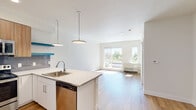 Located at 6190 W 13th Ave in Lakewood, Colorado, this single-floor unit showcases a contemporary design with a clean, neutral palette throughout. The open-concept layout seamlessly connects the kitchen and living area, creating a bright and functional living space bathed in natural light from large sliding glass doors and windows that offer views of the surrounding greenery.

The kitchen is a standout feature, outfitted with stainless steel appliances including a refrigerator, range, and microwave. Cabinetry combines white lower cabinets with warm wood-toned upper cabinets, complemented by a gray tile backsplash. A peninsula with a white countertop and undermount sink provides additional workspace and casual seating, accented by modern pendant lighting. Teal floating shelves add a pop of color to the design.

Light hardwood-style flooring runs throughout the main living areas, lending warmth and continuity to the space. The living area is generously proportioned and leads toward a bedroom that receives natural light through a window with exterior views. The bathroom is equipped with a full soaking tub and shower combination, chrome fixtures, and clean white finishes. An entry door with a decorative patterned design adds a distinctive architectural touch to the interior.