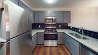 Located at 34 Woodcross Dr in Columbia, SC, this single-floor residence offers a well-designed layout with a consistent, neutral aesthetic throughout. The kitchen is a standout feature, showcasing grey shaker-style cabinetry, granite-look countertops, a dark subway tile backsplash, and stainless steel appliances including a refrigerator, electric range with oven, and microwave. Rich hardwood-style flooring grounds the kitchen and extends into the bathroom areas, adding warmth and durability. A pass-through countertop connects the kitchen to the adjacent dining area, which features plush carpeting and a decorative pendant light. The living area is generously sized, with soft carpet, neutral walls, and a window that allows natural light to fill the room. Multiple bedrooms continue the carpeted flooring and are each equipped with ceiling fans, windows with blinds offering outdoor views of surrounding greenery, and ample wall space. The bathroom includes a wide vanity with grey cabinetry, a large mirror, and a full-size soaking tub with a shower surround, complemented by hardwood-style flooring and chrome fixtures. Textured ceilings and clean white trim are consistent throughout, contributing to the home's cohesive and practical interior design.