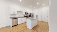 This single-floor residence showcases a clean, contemporary design throughout. The open-concept kitchen is a standout feature, equipped with white shaker-style cabinetry, granite countertops, and a full suite of stainless steel GE appliances, including a gas range, over-the-range microwave, side-by-side refrigerator with ice and water dispenser, and a dishwasher. A central island with matching granite provides additional prep space and casual seating. Light hardwood flooring runs throughout the main living areas, complementing the bright white walls and recessed lighting that create a clean, airy atmosphere.

The bathroom features a white vanity with granite countertop, an undermount sink, and a large mirror, maintaining the cohesive modern aesthetic found throughout the unit. The bedroom offers plush carpeting, a ceiling fan, and a window fitted with blinds that allows natural light to filter in. A dedicated hallway area houses a stacked washer and dryer unit, providing convenient in-unit laundry. A second bathroom is accessible from the hallway. The overall layout is compact yet efficiently designed, with consistent finishes tying each space together seamlessly.