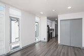 This contemporary single-floor unit features a clean, modern design with a bright and open layout throughout. Wide-plank hardwood-style flooring runs continuously across all areas, providing a warm and cohesive aesthetic that complements the crisp white walls and recessed lighting overhead.

The main living area is generously proportioned and filled with natural light, thanks to floor-to-ceiling windows and a sliding glass door that opens to a balcony with views of the surrounding neighborhood. A transom window above the sliding door further enhances the brightness of the space, creating an airy and welcoming atmosphere.

The kitchen is efficiently designed and positioned along one wall, outfitted with stainless steel appliances including a refrigerator, range, and microwave. White upper cabinetry contrasts with dark lower cabinetry, complemented by a tile backsplash and a single-basin sink. A tall window adjacent to the kitchen brings in additional natural light.

A sliding barn-style door provides access to what appears to be a bathroom or utility area, while a set of bi-fold closet doors offers generous built-in storage. The unit's layout flows intuitively from the living space through to the kitchen and entry corridor, making efficient use of every square foot. Overall, the space presents a polished, move-in-ready aesthetic suited to modern living.
