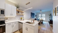 Located at 5395 Martway St in Mission, KS, this thoughtfully designed one-bedroom, one-bathroom residence offers a efficient and stylish single-floor layout. The open-concept kitchen and living area serves as the heart of the home, featuring white shaker cabinetry with black hardware, quartz countertops, a subway tile backsplash, and a full suite of stainless steel appliances including a refrigerator, range, over-the-range microwave, and dishwasher. Warm-toned floating wood shelves add an inviting, modern touch. A peninsula with bar seating provides an ideal casual dining space beneath a sleek linear pendant light. Light hardwood flooring flows seamlessly throughout the main living areas, complementing the neutral color palette. The living room is bright and airy, benefiting from large windows and a ceiling fan, with a glass door opening directly to a private outdoor patio. The bedroom is a calm retreat with plush carpeting, a window with outdoor views, and gold-accented wall sconces. The bathroom features a white vanity with ample storage and a full shower. A dedicated closet completes this well-appointed residence.