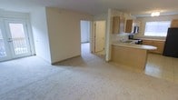 This single-floor space features an open-concept layout that seamlessly connects the living area to the kitchen. The living space is finished with light-colored carpeting that extends throughout, lending a clean and neutral aesthetic. Walls are painted in a soft off-white tone, creating a bright and airy atmosphere that feels open and easy to personalize.

The kitchen is positioned adjacent to the living area, separated by a partial wall with a built-in breakfast bar counter, offering a natural transition between spaces. Medium-toned wood cabinetry lines the upper walls of the kitchen, providing ample storage. The kitchen is equipped with a sink, range, and built-in microwave, making it a fully functional cooking space. Tile flooring in the kitchen contrasts subtly with the carpeted living area.

A doorway visible from the main living space leads to an additional room, suggesting further functional areas within the unit. The structural column near the kitchen acts as a subtle divider without fully closing off the space. Overall, the unit presents a straightforward, functional layout with a clean, move-in-ready feel.