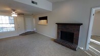 This single-floor space centers around a classic brick fireplace with a dark wood mantel, serving as the focal point of the main living area. The fireplace is constructed with rich, dark-toned brick and features a raised hearth, adding both warmth and character to the room. The living area is finished with neutral-toned carpet, contributing to a comfortable and understated aesthetic throughout.

The ceiling rises notably above the main living space, with an upper clerestory section that adds visual height and an open, airy quality to the room. Ventilation is handled via ceiling-mounted HVAC registers. The walls are finished in a light, neutral tone, keeping the space feeling bright and versatile.

A pass-through window opens from the living area into what appears to be a kitchen or adjacent utility space, allowing for convenient communication between rooms while maintaining defined zones. A hallway visible in the background leads to additional areas of the unit, including a door that likely accesses a bedroom, bathroom, or exterior. The flooring transitions from carpet in the main living space to a harder surface in the connecting hallway.