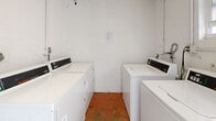 This dedicated laundry room at 3300 El Jardin Dr in Hollywood, Florida, offers a practical and functional space designed exclusively for laundry needs. The room is compact yet efficiently laid out, housing multiple commercial-grade Speed Queen washers and dryers arranged along the walls, maximizing the available floor space. The machines feature digital control panels, reflecting their commercial-grade capability and reliability. The interior is finished with white-painted textured walls and a terracotta tile floor, giving the space a straightforward, utilitarian character. A small window on one wall allows for a degree of natural light and ventilation, contributing to a brighter atmosphere. Exposed plumbing and utility connections are visible along the walls, consistent with the functional nature of the space. Access is provided through a solid wood door. The overall layout is simple and purpose-driven, making it easy to navigate. This laundry room serves as a no-frills, dependable facility well-suited for shared or multi-unit residential use in the Hollywood, FL area.