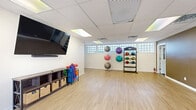 This single-floor fitness studio offers a thoughtfully designed open-plan layout, providing ample room for a variety of exercise activities. The space features light wood-look flooring throughout, which lends a clean and inviting aesthetic while remaining durable for high-activity use. A drop tile ceiling with recessed panel lighting ensures bright, even illumination across the entire floor area.

The studio is divided into functional zones. One area houses a pair of stationary cycling bikes positioned in front of large windows, which flood the space with natural light and offer views of the surrounding outdoor greenery — creating an energizing environment for cardio workouts. An exit door provides convenient direct outdoor access from this section.

On the opposite side of the studio, the open floor area is well-suited for group fitness classes, stretching, or mat-based exercises. A rolling rack holds an assortment of colorful exercise balls in various sizes, while a freestanding medicine ball storage rack offers additional equipment organization. A wall-mounted flat-screen television serves as a focal point for guided workouts or instructional content. A low storage console with basket drawers keeps yoga mats and accessories neatly arranged. Glass block windows along one wall allow natural light to filter in while maintaining privacy. The neutral wall tones and clean, uncluttered layout make this studio a versatile and functional fitness environment.