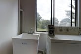 This single-floor utility space is designed with function and practicality at its core. The laundry room is equipped with a deep white utility sink fitted with a chrome faucet, ideal for heavy-duty washing tasks. Adjacent to the sink is a coin-operated washing machine, making this space well-suited for a shared or multi-use facility setting. Exposed plumbing pipes run along the neutral-toned walls, reflecting the room's utilitarian character. A large, dark-framed window dominates one wall, flooding the space with natural light and offering an open view of lush, mature trees and green surroundings outside — creating a pleasant, airy atmosphere despite the room's functional purpose. The clean, light-colored walls and bright natural light contribute to a tidy and well-maintained environment. The layout is straightforward and efficient, with equipment and fixtures arranged to maximize usability within the space. Overall, this laundry facility provides a no-frills, practical environment that prioritizes ease of use, accessibility, and functionality for its users.