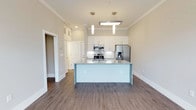 This single-floor residence in Overbrook showcases a thoughtfully designed open-concept layout that seamlessly integrates the kitchen and living areas. The kitchen serves as the focal point of the space, anchored by a substantial center island topped with speckled granite countertops that provide ample workspace and casual dining potential. Sleek white cabinetry extends to the ceiling, maximizing storage while contributing to the bright, airy atmosphere of the space. Stainless steel appliances, including a French door refrigerator with an ice and water dispenser, a range, and a built-in microwave, lend a contemporary and functional edge to the kitchen. Two modern pendant lights hang above the island, complemented by a flush ceiling fixture that ensures thorough illumination throughout. Crown molding lines the ceiling, adding an elegant architectural detail to the otherwise clean and minimalist design. Warm-toned hardwood-style flooring flows throughout, grounding the neutral color palette of soft beige and white walls. The open layout allows natural light to circulate freely, creating a welcoming and functional living environment.