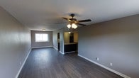 This single-floor space offers a clean, open layout designed for functional everyday living. The main living area is finished with dark hardwood-style flooring that runs throughout, providing a sleek and cohesive look underfoot. Neutral gray walls create a versatile backdrop that complements a variety of décor styles. A ceiling fan with an integrated light fixture is centrally positioned overhead, promoting airflow and providing ambient lighting across the space.

A window along one wall allows natural light to filter into the living area, brightening the interior and creating a welcoming atmosphere. The space flows seamlessly into the kitchen, which is partially separated by a raised breakfast bar counter finished in dark tones, adding definition between the two areas while maintaining an open feel. The kitchen is equipped with dark cabinetry and includes a refrigerator, with under-cabinet lighting that highlights the workspace.

A hallway visible from the main living area suggests additional rooms beyond, and a ceiling-mounted HVAC vent indicates a central air system. White baseboards provide a clean contrast against the dark flooring throughout, adding a polished finishing detail. Overall, the space presents a practical and modern interior with consistent finishes and a straightforward, open design.