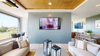 Located atop a building in Fort Worth, TX, this rooftop lounge offers a generously sized, single-floor open-plan space that seamlessly blends indoor comfort with elevated outdoor access. The interior is defined by striking warm-toned wood plank ceilings that add richness and texture, complemented by light wood-look tile flooring throughout. The living area is anchored by a sleek accent wall in a muted blue-gray tone, upon which a large wall-mounted flat-screen television is centered, flanked by plush white and neutral-toned seating arrangements ideal for lounging and socializing. Decorative lantern floor fixtures add an understated elegance to the space. Large floor-to-ceiling windows flood the interior with abundant natural light while framing panoramic views of the Fort Worth skyline and surrounding cityscape. An oversized ceiling fan enhances the airy, resort-like atmosphere. The dining area features a built-in leather banquette with a concrete-top table, a wall-mounted television, and white subway tile accents, creating a cozy yet refined nook. A wet bar with dark wood cabinetry and a sink serves as a functional entertaining feature nearby. Access to the outdoor rooftop terrace is provided through glass-framed doors, extending the lounge experience into the open air with additional seating and sweeping views.