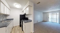 This single-floor unit offers a practical and straightforward layout designed to maximize functionality. The galley-style kitchen is a standout feature, equipped with black appliances including a refrigerator, range, and microwave. White cabinetry lines both sides of the kitchen, providing ample storage, while dark granite-style countertops and a mosaic tile backsplash add a touch of visual interest. Wood-look vinyl flooring runs throughout the kitchen, extending into the entry area.

The living area is open and generously proportioned, featuring neutral carpet and white walls that create a bright, clean atmosphere. A sliding glass door at the far end of the living space allows natural light to filter in and provides access to an outdoor area. The textured ceiling is consistent throughout the unit.

The bedroom is carpeted and comfortably sized, featuring a ceiling fan with light fixture and a window that brings in additional natural light. White walls keep the space feeling open and adaptable.

The bathroom includes a full tub and shower combination with white tile surround, a toilet, and wood-look vinyl flooring, maintaining a clean and cohesive aesthetic. Overall, the unit presents a simple, functional living environment with consistent finishes throughout.