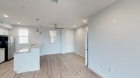 This single-floor space features a clean, contemporary design characterized by light hardwood flooring that extends throughout the open-concept layout. The living and dining area is bathed in natural light from a window fitted with white blinds, contributing to the space's airy and welcoming atmosphere. Recessed lighting in the ceiling is complemented by stylish metallic pendant lights that hang near the kitchen area, adding a modern decorative touch. The kitchen is partially visible and includes a white countertop, maintaining a cohesive, minimalist aesthetic. A ceiling fan is centrally positioned to provide efficient air circulation. The walls are finished in a soft, neutral light blue-gray tone that enhances the sense of openness throughout. A white entry door provides access to the unit, while an additional doorway leads to a separate room or hallway, suggesting a functional and well-organized floor plan. Visible HVAC vents integrated into the ceiling indicate a modern climate control system. Overall, the space presents a fresh, move-in-ready environment with thoughtful design details suited for comfortable everyday living.