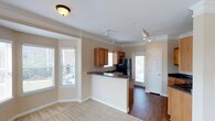 This single-floor residence offers a well-designed open-concept layout that seamlessly connects the living area to the kitchen, creating a cohesive and functional living environment. The living space features plush carpeting and is bathed in natural light from multiple windows fitted with blinds, offering views of the surrounding exterior.

The kitchen is a standout feature, boasting warm honey-toned wood cabinetry and a peninsula with a dark countertop that provides both additional workspace and a natural boundary between the kitchen and living area. A stainless steel refrigerator and black appliances complete the kitchen setup. A window above the sink brings in additional natural light, enhancing the brightness of the cooking space.

Crown molding runs throughout the home, adding a refined architectural detail to the neutral beige walls and white trim. Track lighting and flush-mount ceiling fixtures provide ample illumination throughout. Rich hardwood flooring extends through the kitchen and adjoining hallway, which leads to a door opening to an outdoor area, further expanding the sense of space. The overall ambiance is warm, bright, and inviting, with a clean, classic aesthetic that suits a variety of lifestyles.