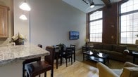 This thoughtfully designed urban space blends industrial character with modern comfort. The open-plan layout seamlessly connects the dining and living areas, creating a cohesive and functional environment. Exposed brick walls add a distinctive, loft-like aesthetic, while the soaring ceilings amplify the sense of spaciousness throughout. Large, multi-pane arched windows are a standout architectural feature, drawing in abundant natural light and offering views of the surrounding cityscape. Light hardwood flooring runs throughout, complementing the warm tones of the brick and neutral wall palette. The dining area features a dark wood table and chairs set alongside a granite-topped breakfast bar with wicker seating, ideal for casual or formal dining. The living area is anchored by a plush sofa, a sleek glass-top coffee table, and a wall-mounted flat-screen television, all arranged to encourage relaxed gathering. A ceiling fan with track lighting provides both comfort and practicality. The overall ambiance strikes a balance between the rugged charm of a historic building conversion and the clean, refined touches of contemporary interior design.