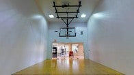 This indoor recreational facility centers around a full basketball court finished with gleaming hardwood flooring, offering a polished and professional playing surface. The space is defined by its impressively tall ceilings, which accommodate a ceiling-mounted basketball hoop and backboard system — a standout architectural feature that maximizes vertical clearance and ensures an authentic playing experience. The crisp white walls reflect the bright overhead lighting provided by recessed and flush-mount ceiling fixtures, creating a well-lit environment suited for active use at any time of day. At the far end of the court, large glass doors and windows open directly into an adjoining fitness center, offering a seamless transition between athletic spaces. The fitness area beyond is visible through the glass, revealing exercise equipment and natural light filtering in from exterior windows. The overall design is clean, functional, and purpose-built, with safety signage and ventilation systems thoughtfully integrated into the walls. Together, the basketball court and connected gym create a comprehensive, amenity-rich athletic environment designed to support a wide range of fitness and recreational activities.