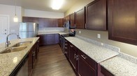 This single-floor unit offers a thoughtfully designed layout that balances functionality and style. The kitchen serves as the centerpiece, showcasing rich dark cabinetry paired with speckled granite countertops and a full suite of stainless steel appliances, including a refrigerator, range, and built-in microwave. A large central island with an undermount sink and pendant lighting above provides additional workspace and a natural gathering point. The kitchen flows seamlessly into the open living area, which features dark hardwood flooring throughout and neutral-toned walls that create a clean, versatile backdrop. Natural light enters through windows fitted with vertical blinds, brightening the space. The bedroom continues the neutral palette with plush carpeting underfoot, a window with vertical blinds, and baseboard heating for year-round comfort. The bathroom is finished with the same dark hardwood flooring found throughout the unit, complemented by a dark vanity with granite countertop, a full soaking tub with shower, and clean white walls that keep the space feeling fresh and bright. Overall, the unit presents a cohesive and contemporary aesthetic with quality finishes carried consistently from room to room.
