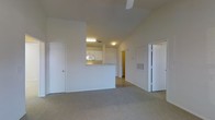 This single-floor space features an open-concept layout that connects the living area to the kitchen via a half-wall pass-through counter, creating a sense of openness while maintaining a defined cooking zone. The living area is finished with wall-to-wall carpet in a neutral tone, contributing to a comfortable and cohesive aesthetic throughout.

The kitchen is positioned toward the center of the unit, equipped with white cabinetry and a microwave visible above the counter. A ceiling-mounted light fixture illuminates the kitchen area, while a flush-mount light provides additional brightness in the adjacent space. The white walls throughout the unit lend a clean, bright atmosphere.

Multiple doors are visible from the main living space—one to the left and one to the right—likely leading to bedrooms, a bathroom, or storage areas. A hallway extends toward the back of the unit, hinting at additional rooms beyond. A wall-mounted HVAC vent and ceiling fan contribute to climate control. Overall, the space offers a functional, straightforward layout suited for everyday living.
