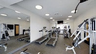 Located at 2131 N. Meridian, this fitness center offers a well-equipped, single-floor exercise facility designed to support a comprehensive workout routine. The space is finished with a clean, modern aesthetic, featuring bright white walls and recessed circular lighting that evenly illuminate the room throughout. Durable rubber flooring covers the exercise area, providing both safety and comfort underfoot. A full-length mirror runs along one wall, enhancing the sense of openness while allowing users to monitor their form during workouts. The cardio section is well-stocked with multiple treadmills and additional aerobic machines, offering ample options for cardiovascular training. Strength and resistance equipment is also present, rounding out the facility's capabilities. A wall-mounted flat-screen television is positioned for easy viewing from the cardio equipment, adding an entertainment element to workout sessions. Natural light filters in through a window at the far end of the room, contributing to the bright and energizing atmosphere. The layout is thoughtfully organized, allowing for comfortable movement between stations. Overall, this fitness center provides a functional, well-maintained environment suited for a variety of fitness levels and training needs.
