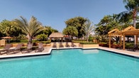 This single-story property showcases a standout outdoor amenity area centered around a generously sized swimming pool with clear, turquoise water. The pool deck is finished in light-toned pavement and lined with dark-framed lounge chairs, providing ample space for relaxation and sunbathing. A thatched-roof palapa structure serves as a shaded gathering spot, lending a tropical, resort-inspired character to the outdoor space. The surrounding landscape is richly planted with mature palm trees, tropical shrubs, and colorful flowering plants, creating a lush, private atmosphere. Warm-toned stucco walls and planter borders frame the perimeter, contributing to the cohesive, tropical aesthetic. The area benefits from abundant natural light and open sky views, enhancing the outdoor living experience. The remaining interior images were too dark to yield usable detail, so interior features of the property cannot be described at this time. Overall, the outdoor environment presents a well-maintained, resort-style setting well-suited for warm-weather living and outdoor entertaining.