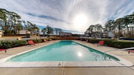 Located at 5100 Highpoint Rd in Union City, Georgia, this outdoor amenity space centers around a generously sized rectangular swimming pool set within a well-defined pool deck. The pool area is enclosed by a sturdy iron security fence, providing a secure and private environment for residents. The surrounding concrete sundeck offers ample space for relaxation, furnished with vibrant red lounge chairs positioned along the perimeter of the pool.

A gabled clubhouse or amenity building is situated directly adjacent to the pool area, featuring large windows and a welcoming entrance, and serves as a functional support structure for the pool facility. A covered pergola-style structure adds an additional shaded gathering spot near the pool deck.

Manicured hedges and well-maintained landscaping line the pool enclosure, contributing to a clean and organized aesthetic. Tall pine trees form a natural backdrop that surrounds the entire area, offering a sense of seclusion and a connection to the natural environment. The open-sky setting allows for abundant natural light throughout the day.

The pool itself is clearly marked with depth indicators and a "No Diving" sign, reflecting attention to safety. The overall layout is thoughtfully designed to serve as a community leisure destination, balancing functionality with an inviting outdoor atmosphere ideal for warm Georgia days.