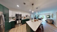 This thoughtfully designed single-floor residence offers a seamless blend of modern aesthetics and functional living. The centerpiece of the space is a striking open-concept kitchen, distinguished by bold forest green lower cabinetry paired with crisp white upper cabinets and light-toned quartz countertops. A generously sized island with bar seating serves as a natural gathering point, illuminated by elegant pendant lights. Stainless steel appliances, including a French door refrigerator, electric range, microwave, and dishwasher, complete the well-appointed cooking space. Light wood-look flooring flows throughout, unifying the kitchen with the adjacent living and dining areas. The living room is bright and welcoming, benefiting from natural light and direct access to a private balcony overlooking lush greenery. The bedroom is a calm retreat, featuring a large window that frames outdoor views and creates a serene atmosphere. The bathroom is finished with slate blue cabinetry, black hardware accents, and wood-look tile flooring, offering both style and practicality with ample cabinet storage. Neutral wall tones and recessed lighting throughout the residence contribute to a clean, contemporary ambiance that feels both polished and livable.