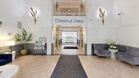 The lobby of Chestnut Oaks, located at 1800 Palmer Rd in Fort Washington, MD, offers a polished and welcoming first impression across a single floor. The entrance hallway is defined by its grand double doors crowned with an arched transom window, flanked by large side windows that flood the space with abundant natural light. Built-in bench seating lines both sides of the entry vestibule, providing a practical and refined welcome. Light wood herringbone-patterned flooring and a central runner guide visitors from the entrance toward the elevator bay, which is framed by a decorative overhead balcony bearing the Chestnut Oaks signage. Gold leaf wall sconces and botanical wall art add warmth and a refined character throughout. The adjoining living area features dramatically soaring ceilings with white paneled walls accented by a two-tone treatment in muted sage green below the chair rail. The space is furnished with a rich navy velvet sofa, a round marble-top coffee table, and a pair of plush gray accent chairs arranged around a wall-mounted flat-screen television. Curated artwork and decorative accents complete the sophisticated aesthetic. Together, the hallway and living room create a cohesive, light-filled lobby that balances elegance with comfort.
