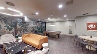 Located on Wilshire Boulevard in the heart of Los Angeles, this amenity lounge offers a well-appointed recreational space designed for both socializing and entertainment. The single-level layout is anchored by a full-size billiards table with a blue felt surface, positioned beneath a rectangular pendant light and accompanied by a mounted cue rack along the wall. A bold tropical botanical mural serves as a dramatic focal point, adding vibrant contrast to the otherwise neutral sage-green walls and dark tile flooring that runs throughout the space.

The lounge area is furnished with a tufted caramel leather sofa, modern curved accent chairs, a black marble coffee table, and sculptural white side tables, all arranged beneath a circular halo-style pendant light. A wall-mounted flat-screen television is positioned above a scalloped wood credenza, creating a comfortable media viewing setup. A tall potted palm adds a natural, organic element to the corner.

A separate dining area features a round pedestal table surrounded by upholstered chairs, ideal for casual gatherings. Bar-height seating is also visible along one wall. Recessed lighting throughout ensures even, ambient illumination, contributing to the space's refined yet relaxed atmosphere.